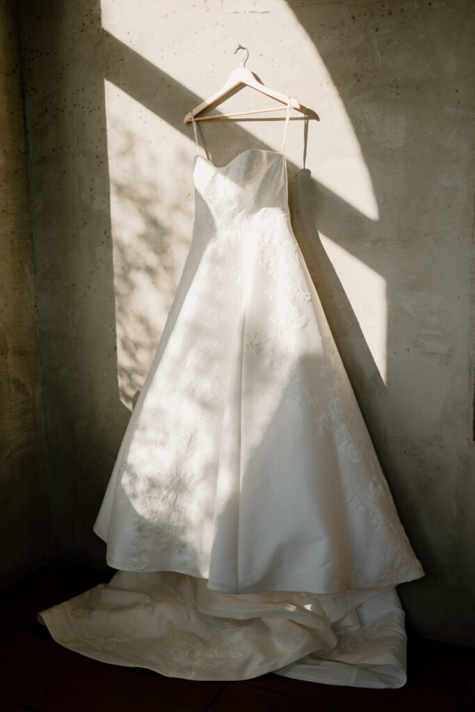 strapless long wedding gown hanging on a wall with the sun reflecting on the beautiful dress