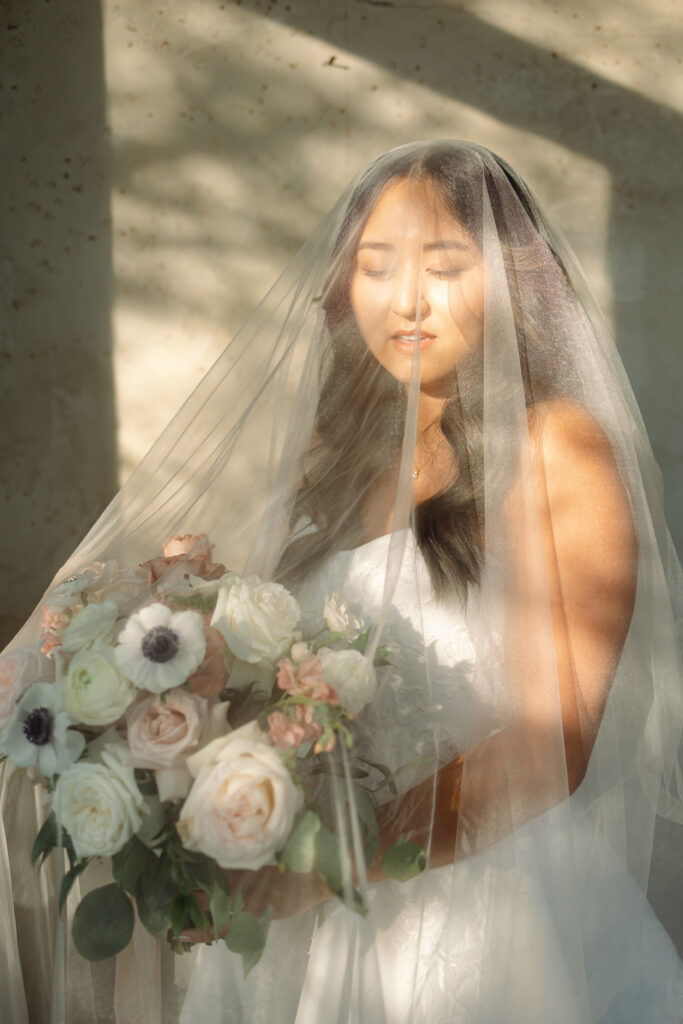 bride holding flower bouquet while veil is covering her face and standing in hallway with large windows and tons of natural light