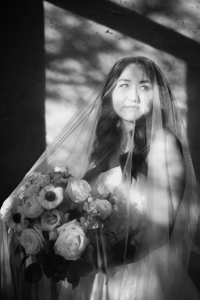 bride holding flower bouquet while veil is covering her face and standing in hallway with large windows and tons of natural light