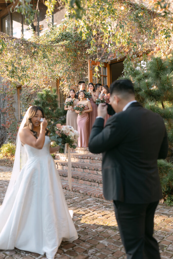 groom and bride wiping tears from eyes after looking at bride for first time during first look wedding photos with their wedding party looking at them from behind