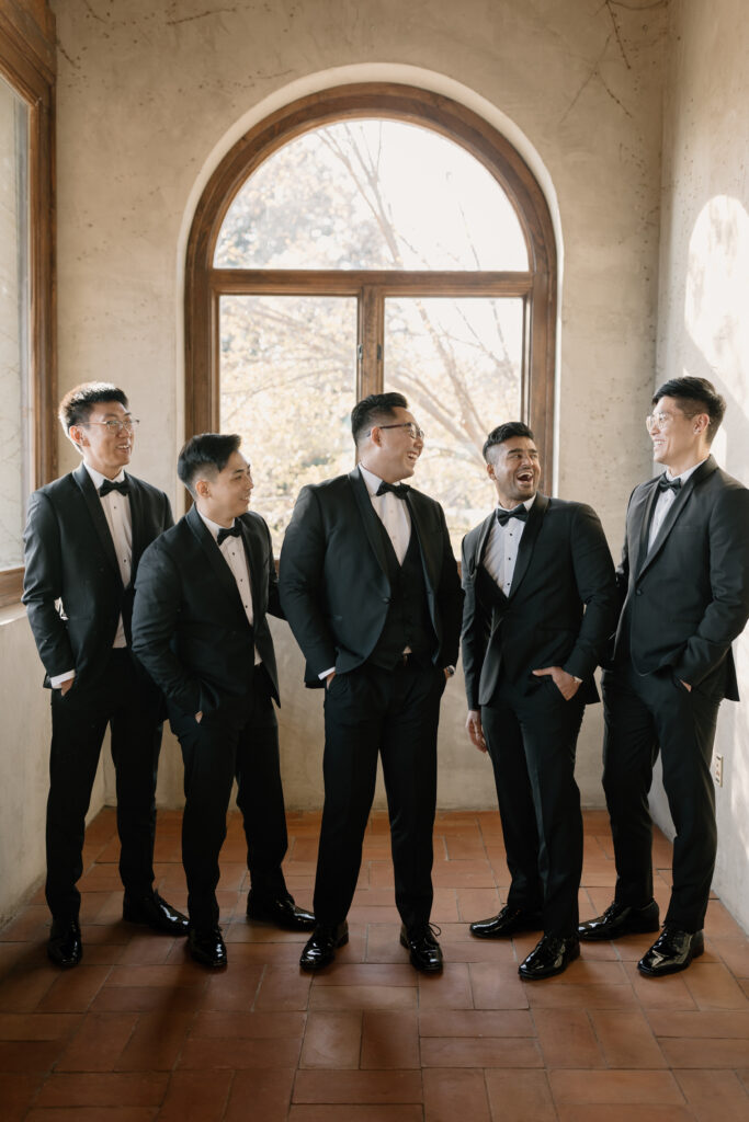 groom and groomsmen standing and laughing at each other while posing in front of window