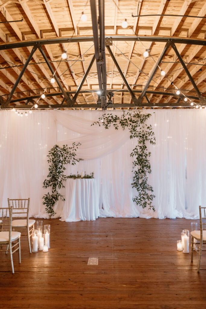 modern wedding venue with gold and white chairs in front of simple wedding arch with candles at the bottom of chairs
