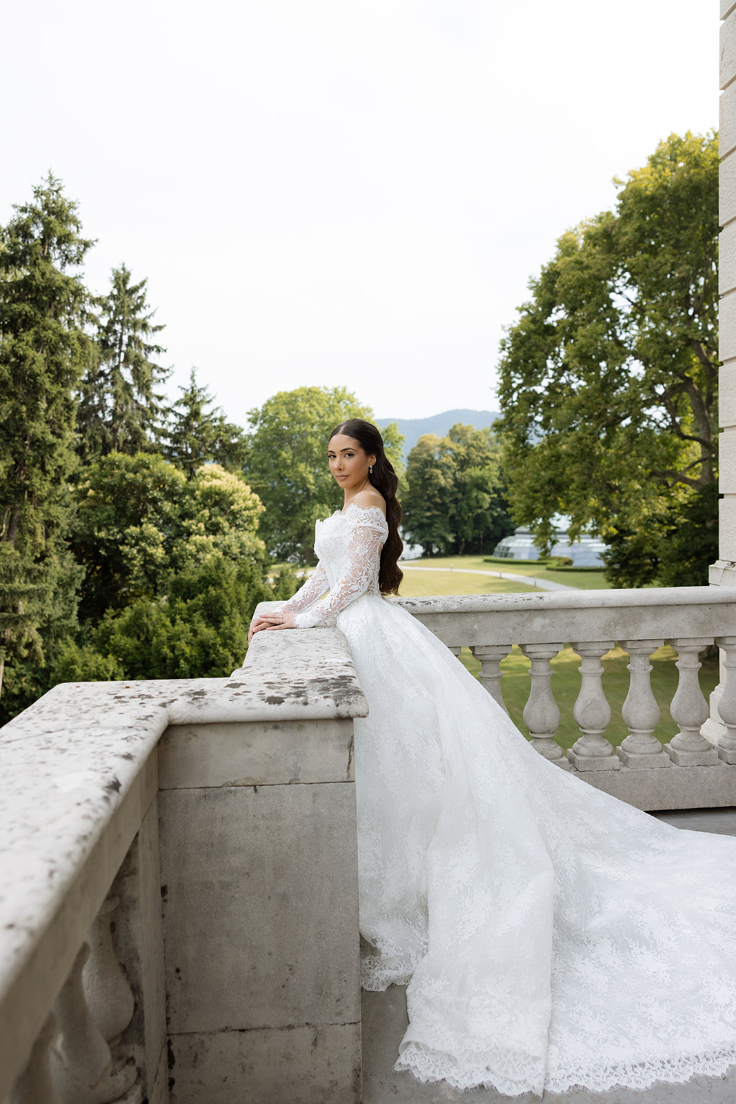 Bride in off-shoulder lace ballgown with cathedral train seated on stone balustrade overlooking Villa Erba's formal symmetrical gardens