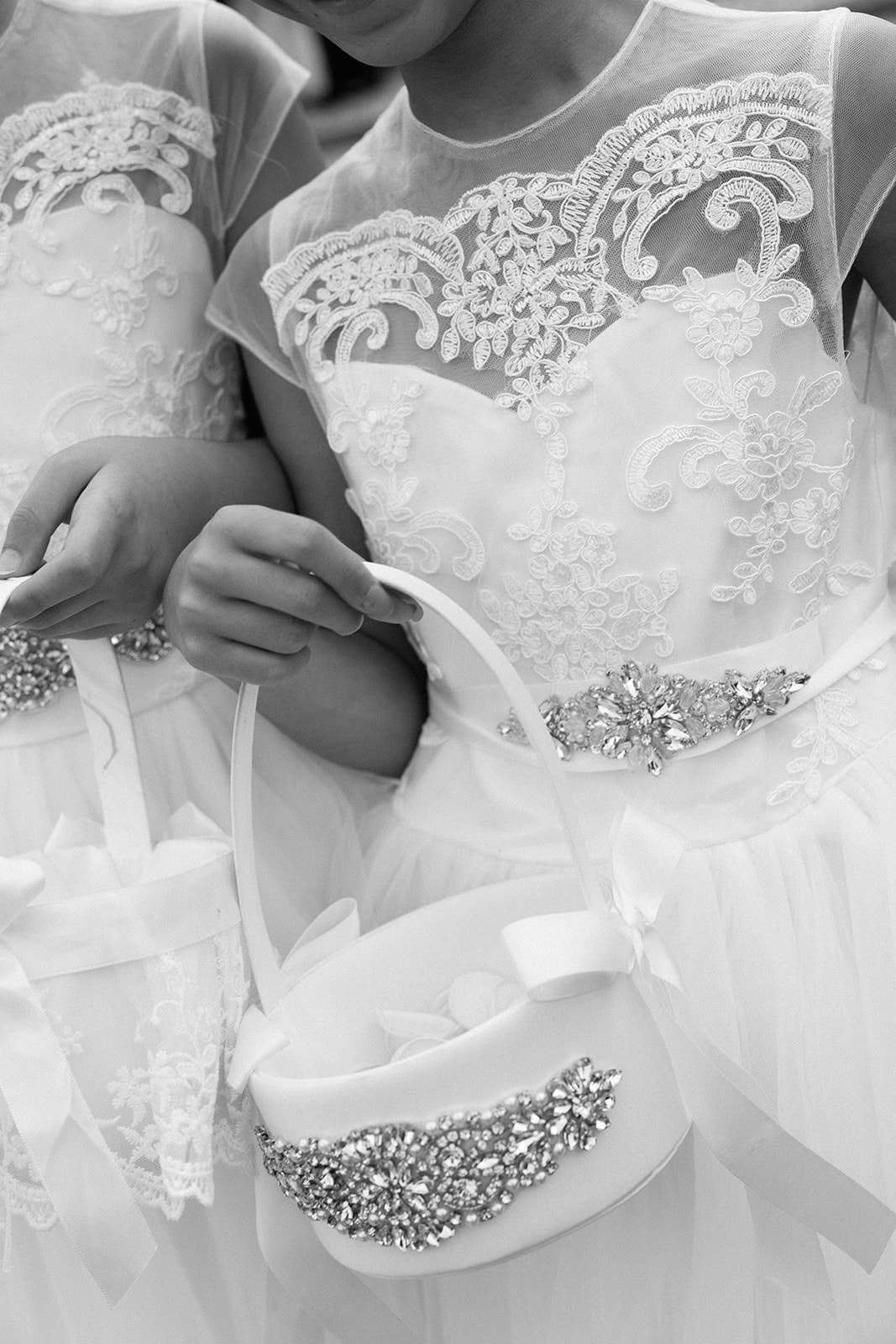 white close-up of flower girls in embroidered lace dresses with crystal sash embellishments, holding matching crystal-buckled flower baskets
