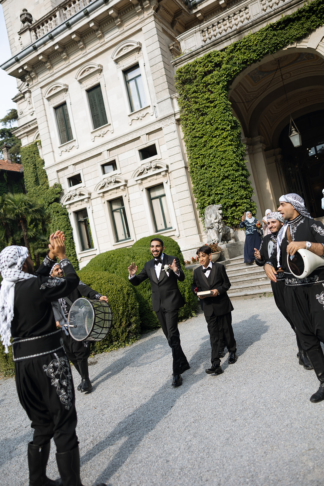 Groom dancing through a traditional Middle Eastern musical procession with drummers and musicians in embroidered attire outside the ivy-covered entrance of Villa Erba