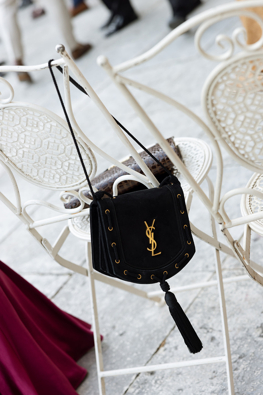 Black Saint Laurent suede bag with gold monogram hardware and tassel resting on a white ornate iron garden chair at the wedding ceremony