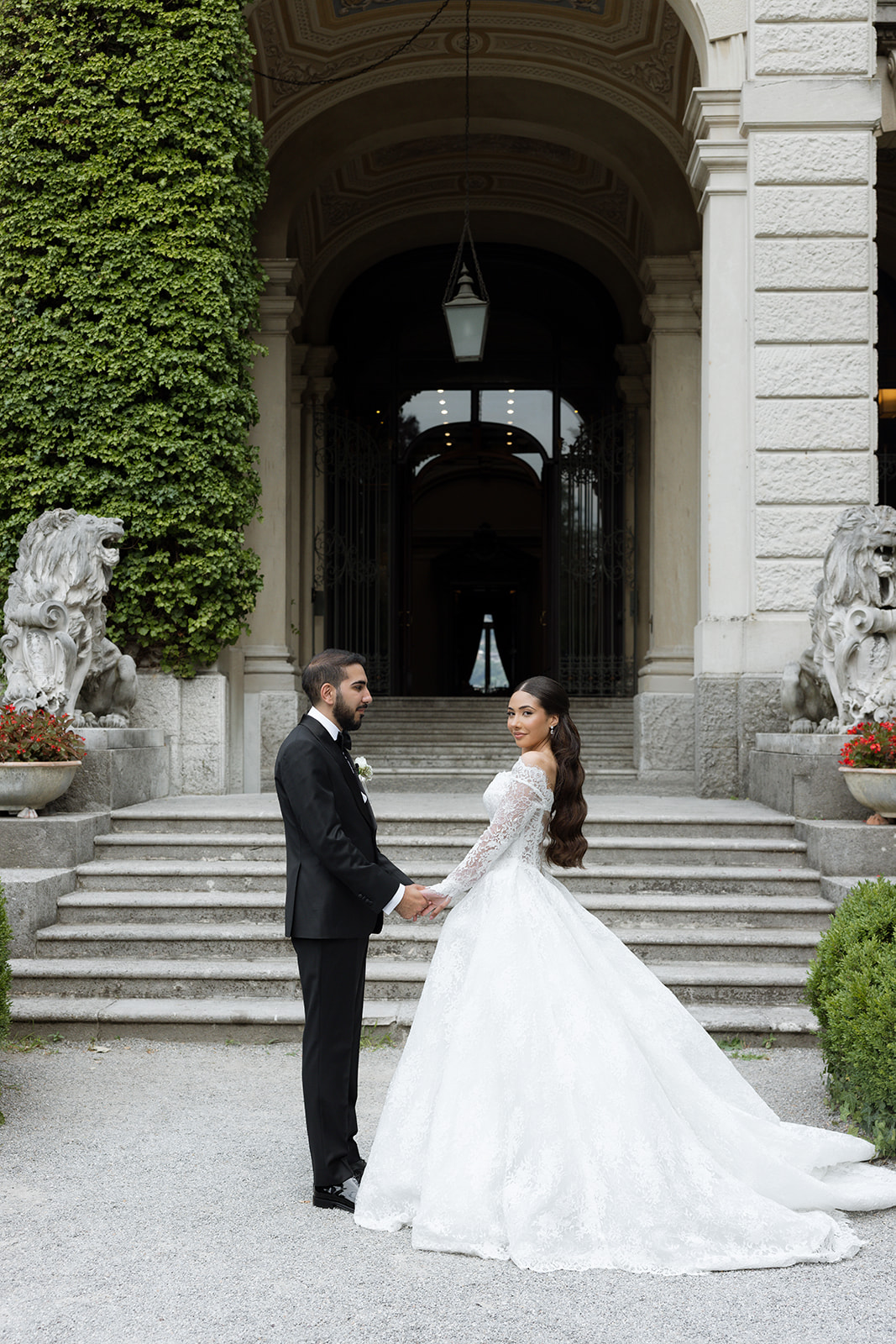 Bride and groom holding hands and turning back toward the camera in front of Villa Erba's ivy-covered grand arched entrance, stone lion statues flanking the steps and the bride's lace cathedral train spread across the gravel