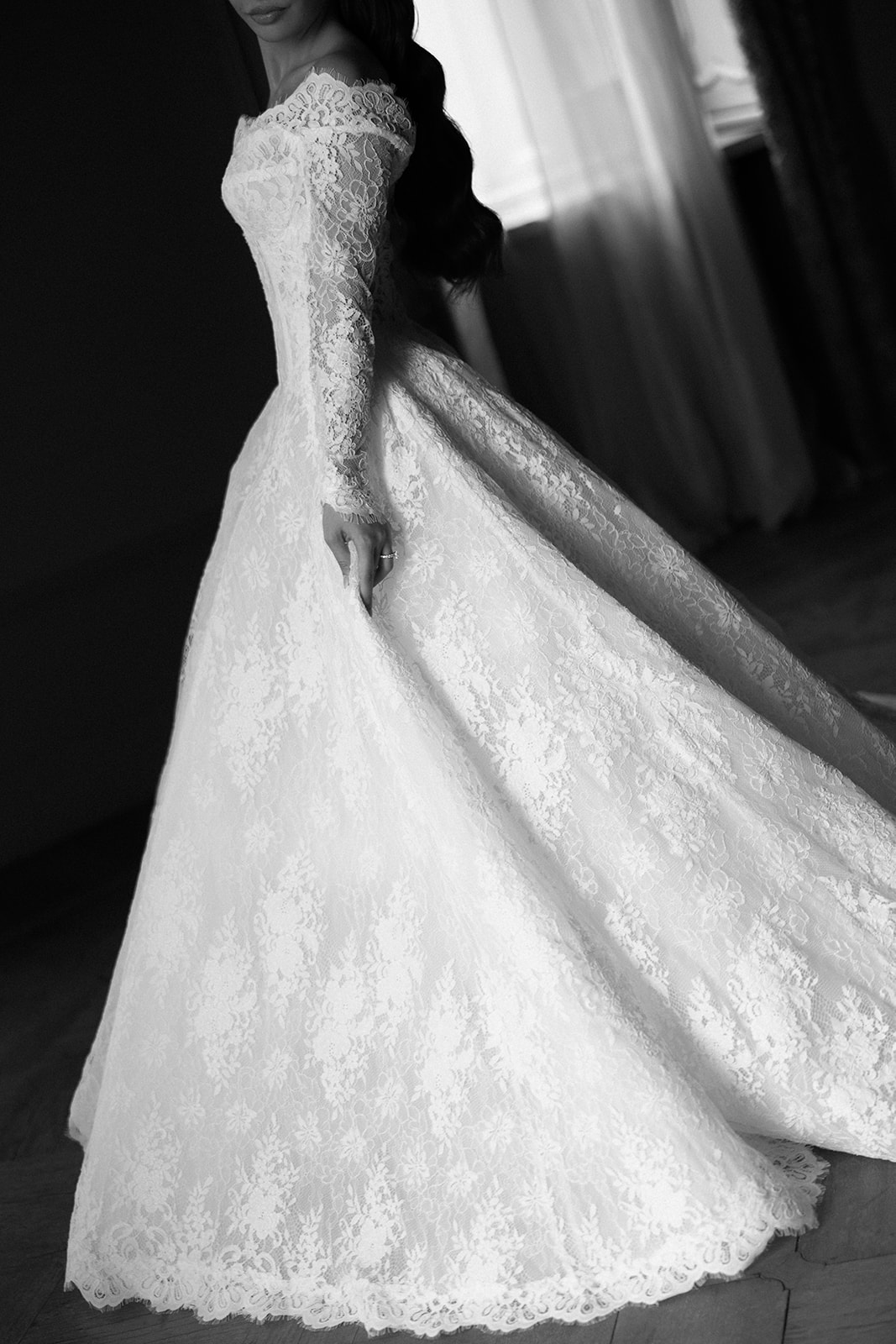 Black and white close-up of bride lifting the full skirt of her lace ballgown, showcasing the intricate floral lace pattern across the fabric and long fitted sleeve