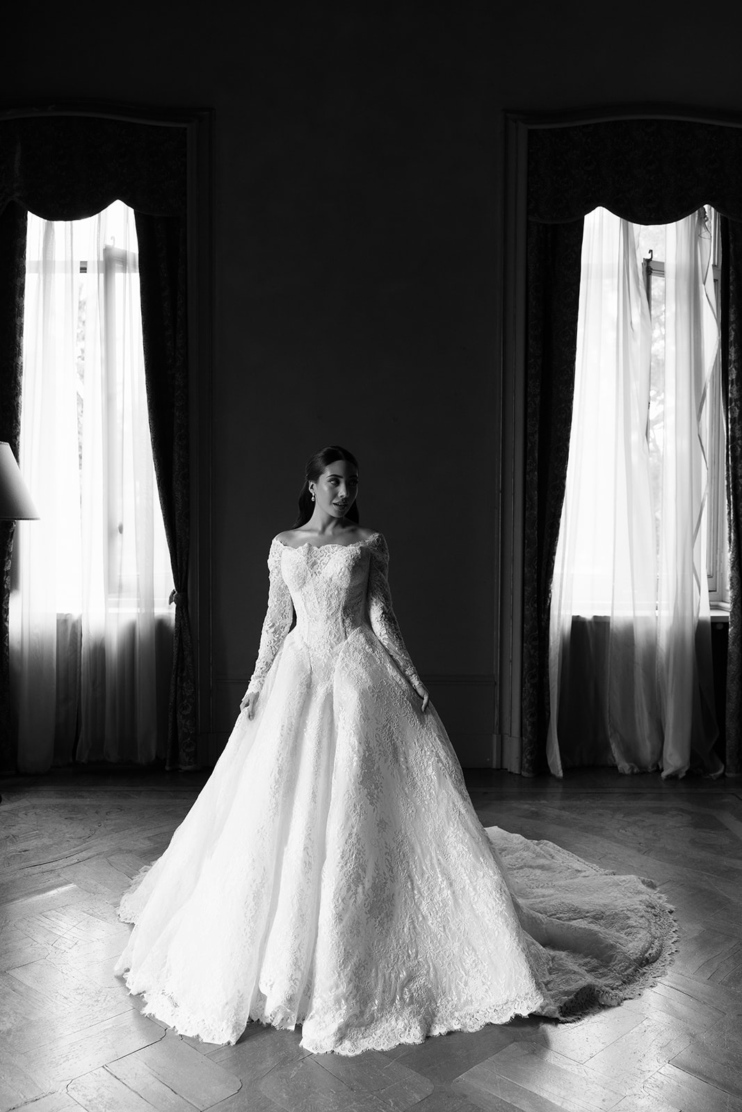 Black and white full-length portrait of bride in off-shoulder lace ballgown with cathedral train, standing between two tall windows in an ornate dim room at Villa Erba, light streaming in from both sides