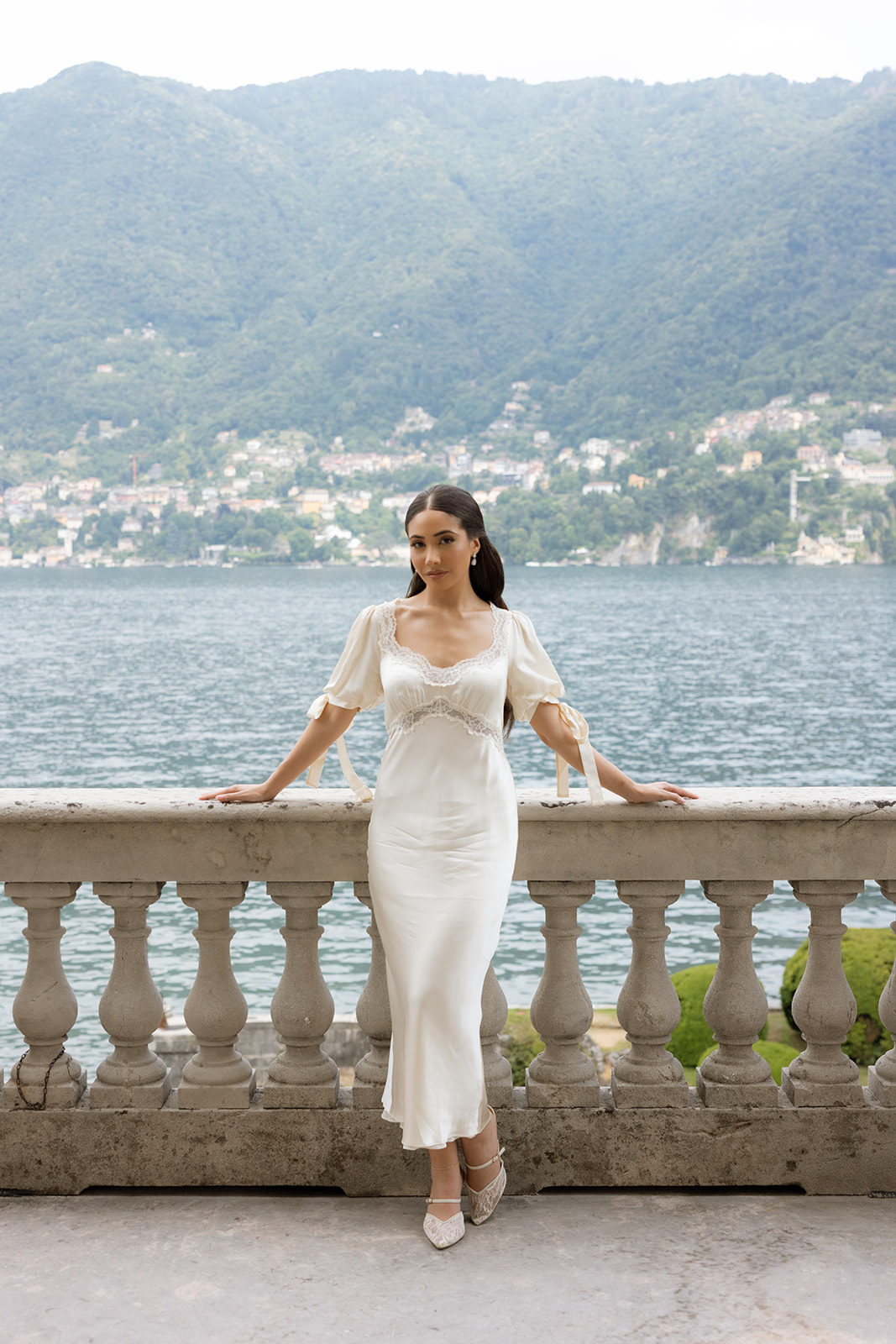 Bride in an ivory silk slip dress with lace trim and puff sleeves posing on the Villa Erba stone balustrade for Lake Como wedding portraits, the Alps and blue water stretching behind her