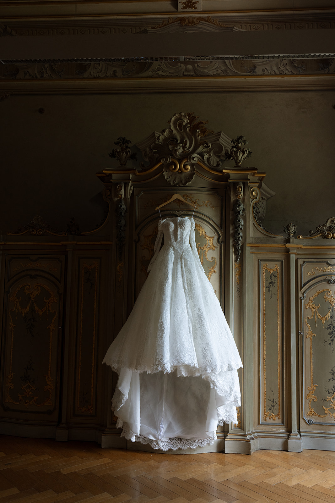 Bride's off-shoulder lace ballgown hanging from a wooden hanger in front of an ornate gilded Baroque armoire with painted panels and gold carved moldings in a Villa Erba suite