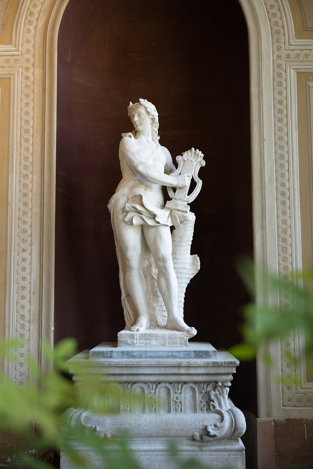 White marble classical statue of a figure holding a lyre, set in an ornate arched niche with decorative column moldings inside Villa Erba's interior galleries, framed by green foliage in the foreground
