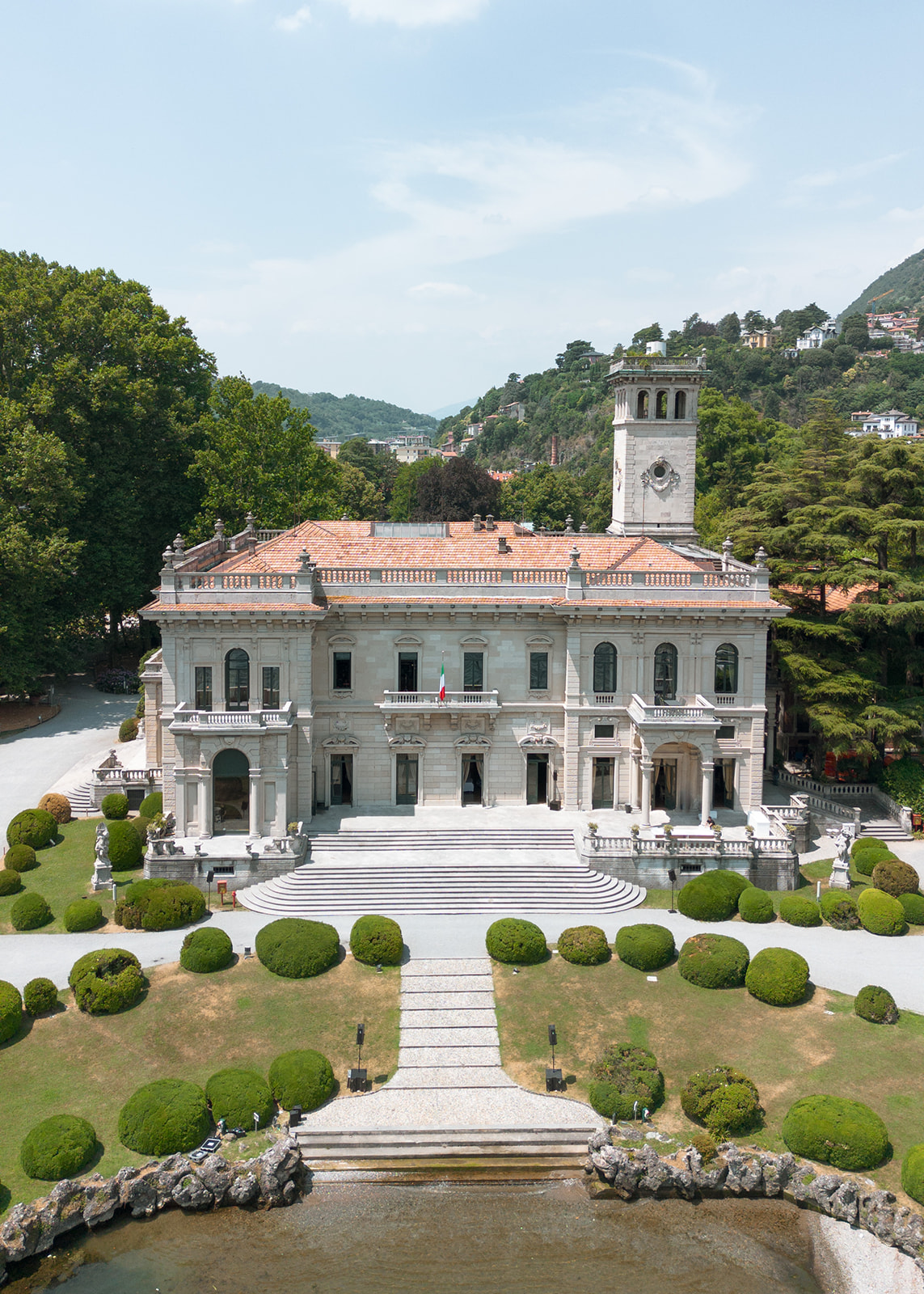 Aerial drone view of Villa Erba's neoclassical main facade in Cernobbio, showing the symmetrical formal garden with ball topiary, grand curved entrance steps, the estate clock tower, and the Italian flag, a defining Lake Como wedding venue