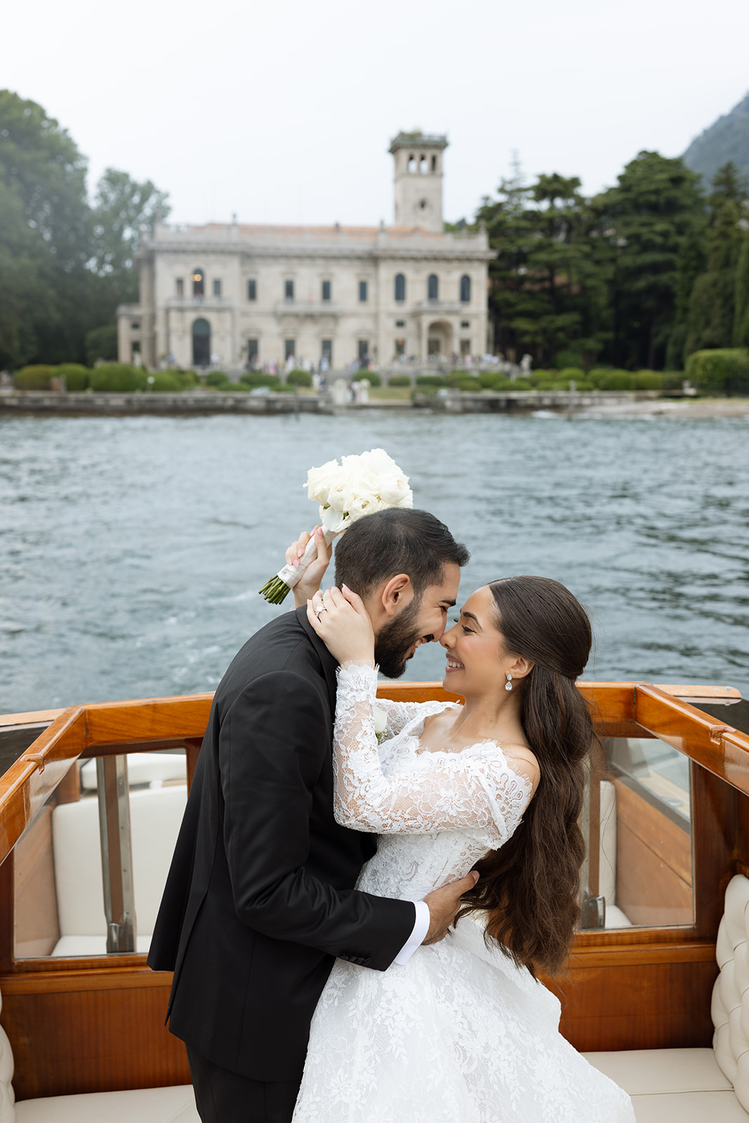 Bride and groom sharing a tender moment on a vintage wooden boat during Lake Como wedding portraits, Villa Erba estate visible across the water
