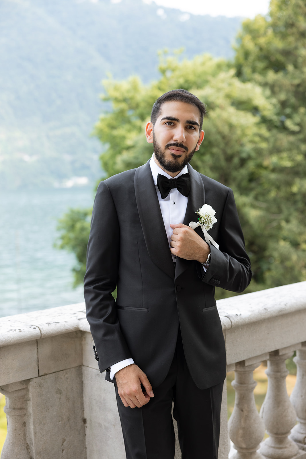 Groom in black tuxedo with white boutonniere adjusting his jacket on the stone balustrade terrace of Villa Erba overlooking the lake