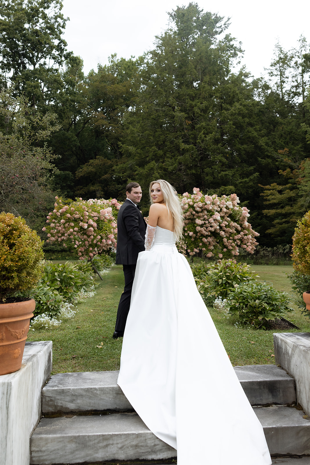 Bride and groom glance back at the camera while pausing on stone garden steps, lush pink hydrangea bushes filling the formal Berkshire estate garden behind them