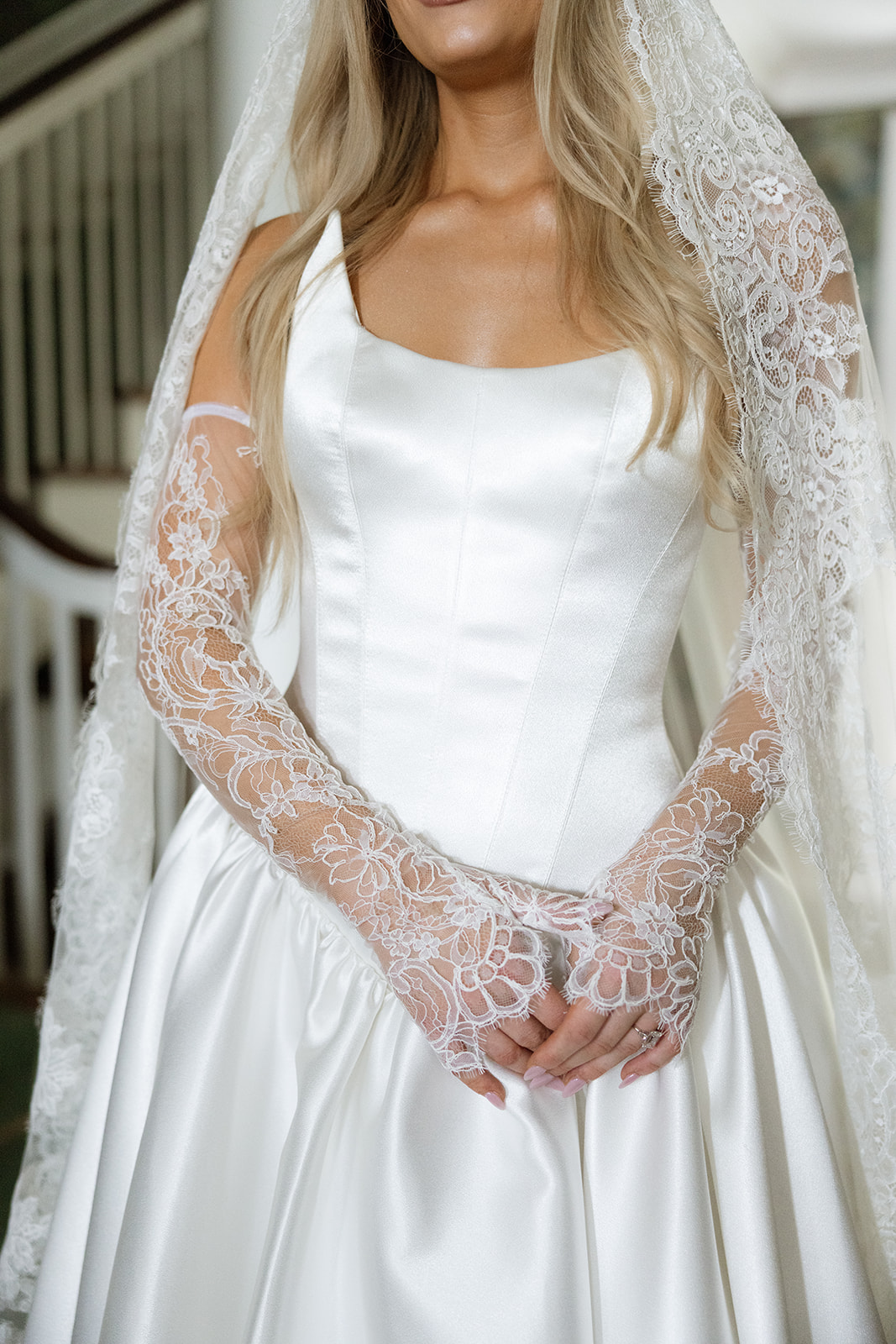 Bride in white satin corset bodice with intricate lace gloves, ornate staircase railing softly blurred in the background