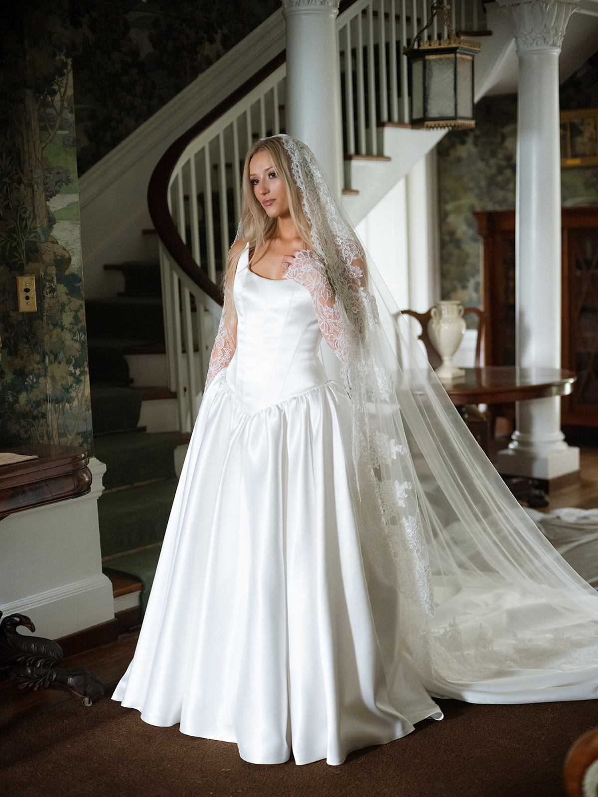 Bride in a flowing white gown with a long lace veil at Chesterwood Stockbridge MA, posed in front of a curved grand staircase framed by botanical wallpaper and ornate columns